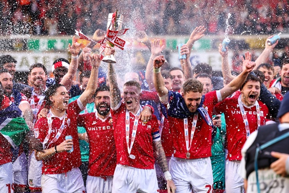 ames McClean of Wrexham lifts the trophy with team mates after winning promotion during the Sky Bet League One match between Wrexham AFC and Charlton Athletic FC at Racecourse Ground on April 26, 2025 in Wrexham, Wales.