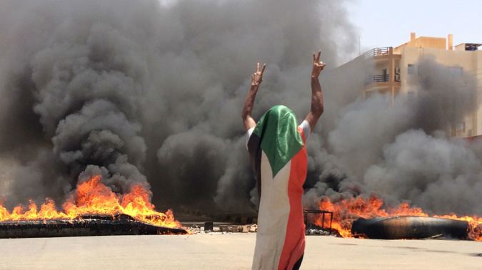 A protester wearing a Sudanese flag flashes the victory sign in front of burning tires and debris on road 60, near Khartoum's army headquarters, in Khartoum. (Photo / AP)