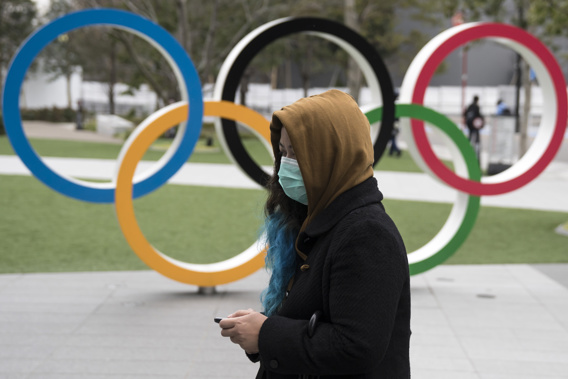 A woman wearing a face mask walks past the Olympic rings in front of the new National Stadium, the main stadium for the upcoming Tokyo 2020 Olympic and Paralympic Games, on February 26, 2020 in Tokyo, Japan.