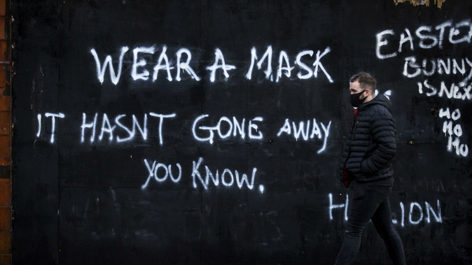A man wearing a face covering walks past graffiti on the Lower Newtownards Road in Belfast with a message reading 'Wear a mask, it hasn't gone away you know'. (Photo / AP)