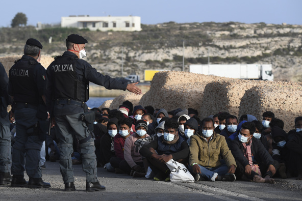 Migrants wearing face masks to curb the spread of COVID-19 sit at a pier as Italian police officers stand by, on the Sicilian island of Lampedusa, southern Italy. (Photo / AP) 