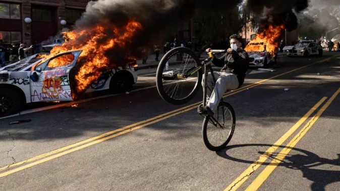 A demonstrator rides a bicycle past burning Waymo vehicles during the Los Angeles protests. Photo / AFP